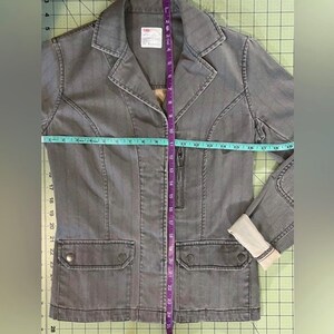 May include: A gray striped denim jacket with a zipper closure and two flap pockets. The jacket is measured with a tape measure, showing the length to be 25 inches.