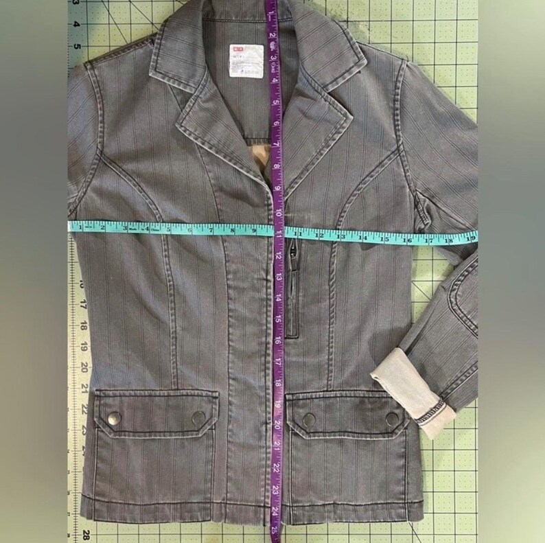 May include: A gray striped denim jacket with a zipper closure and two flap pockets. The jacket is measured with a tape measure, showing the length to be 25 inches.