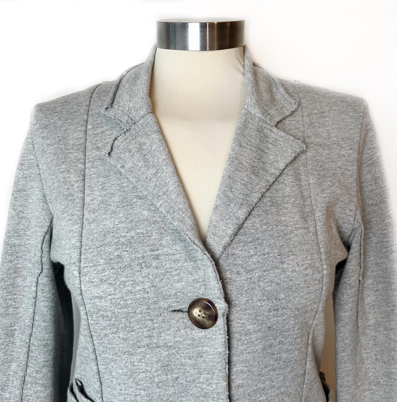 Op de afbeelding: Een grijze blazer met een enkele knoopsluiting. De blazer heeft een reverskraag en lange mouwen.
