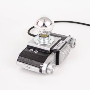 Könnte beinhalten: Eine einzigartige Lampe aus einem Vintage-Kameragehäuse. Die Lampe verfügt über eine silberne Glühbirne und ein schwarzes Netzkabel. Das Kameragehäuse ist schwarz und silber, mit einem klassischen Design. Ein kreativer Einrichtungsgegenstand.