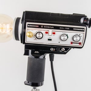 Könnte beinhalten: Schwarze und silberne Vintage-Filmkamera Cosina DXL-755 mit einer großen Edison-Glühbirne. Die Kamera hat mehrere Knöpfe und Schalter, darunter "Back Light", "Auto" und "Slow Motion".