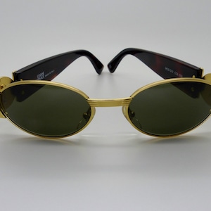 Puede incluir: Gafas de sol con montura dorada y lentes ovaladas verdes. Las patillas tienen un diseño de carey y la montura es dorada. Las gafas tienen el texto "GRW" y "MADE IN ITALY" en la patilla.