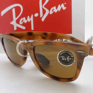 May include: Tortoiseshell-patterned sunglasses with brown lenses. The sunglasses have a classic design with a prominent browline and a white rubber band holding the arms together. The Ray-Ban logo is visible on the lenses and the packaging.