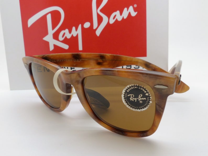 May include: Tortoiseshell-patterned sunglasses with brown lenses. The sunglasses have a classic design with a prominent browline and a white rubber band holding the arms together. The Ray-Ban logo is visible on the lenses and the packaging.