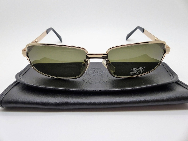 Puede incluir: Gafas de sol con montura dorada y lentes verdes sobre un estuche de cuero negro. Las gafas tienen forma rectangular y patillas negras. El estuche tiene un dise&ntilde;o en relieve y el logotipo de Versace.