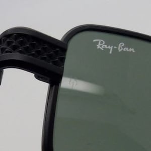 May include: Close-up of black sunglasses with the Ray-Ban logo in white on the lens. The frame is black, and the arm features a geometric pattern. The lenses are a muted green color.