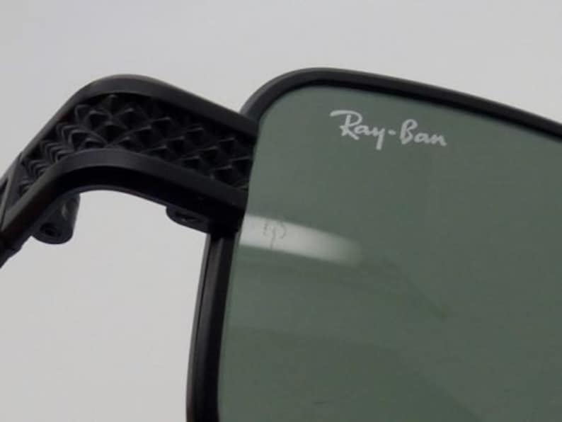 May include: Close-up of black sunglasses with the Ray-Ban logo in white on the lens. The frame is black, and the arm features a geometric pattern. The lenses are a muted green color.