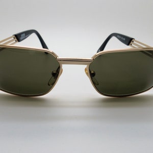 Puede incluir: Gafas de sol doradas con lentes verde oscuro. Las monturas tienen forma rectangular con doble puente y varillas negras. El nombre de la marca es visible en la varilla izquierda.