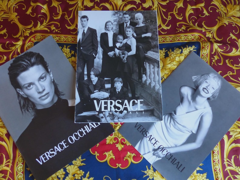 K&ouml;nnte beinhalten: Drei Versace-Modeanzeigen werden auf einem gemusterten Stoff gezeigt. Die zentrale Anzeige zeigt eine Gruppe von Personen. Zwei weitere Anzeigen zeigen einzelne Models. Der Text "VERSACE OCCHIALI" ist auf zwei der Anzeigen zu sehen.