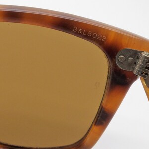 May include: Close-up of vintage sunglasses with a tortoiseshell pattern frame. The lenses are a warm brown color. The text "B&L5022" is visible on the frame. The hinge is made of metal.