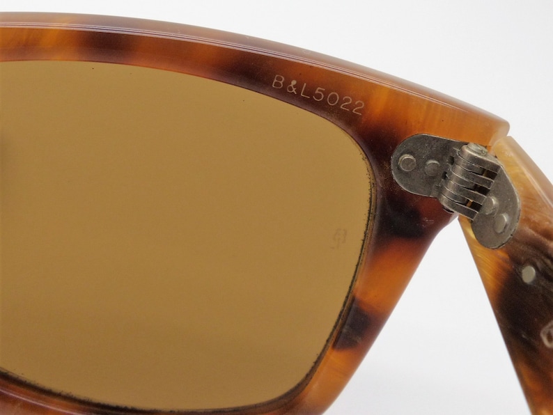 May include: Close-up of vintage sunglasses with a tortoiseshell pattern frame. The lenses are a warm brown color. The text "B&L5022" is visible on the frame. The hinge is made of metal.