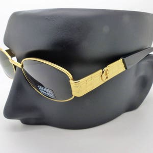 Puede incluir: Gafas de sol con montura dorada y lentes oscuras. Las gafas tienen una montura rectangular dorada con un patrón decorativo en las patillas y una varilla negra. Las lentes son oscuras y las gafas se exhiben en una cabeza de maniquí negra.