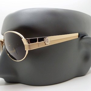 Puede incluir: Gafas de sol doradas con lentes oscuras. Las gafas tienen una montura redonda y una patilla de metal dorado con un emblema decorativo. Las gafas se exhiben en una cabeza de maniquí gris.