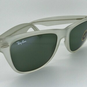 May include: A pair of Ray-Ban sunglasses with a frosted, off-white frame and dark green lenses. The Ray-Ban logo is visible on the left lens. A small sticker is on the right lens. The sunglasses are set against a white background.