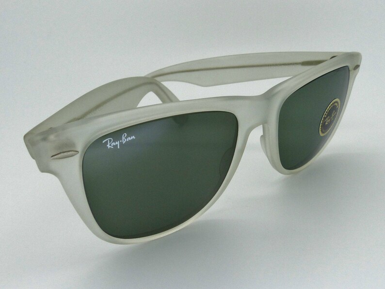 May include: A pair of Ray-Ban sunglasses with a frosted, off-white frame and dark green lenses. The Ray-Ban logo is visible on the left lens. A small sticker is on the right lens. The sunglasses are set against a white background.