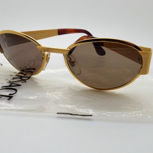 Puede incluir: Gafas de sol doradas con lentes marrones. Las gafas tienen una forma ovalada distintiva y una montura dorada. Las patillas son una mezcla de oro y marrón. Las gafas están sobre una superficie blanca con una bolsa de plástico con la palabra VERSACE impresa.