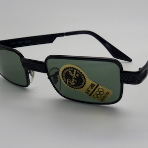 May include: Black Ray-Ban sunglasses with green lenses. The rectangular frames have a textured design on the temples and bridge. The lenses have a sticker with the Ray-Ban logo and Olympic rings. The text "G-15 LENS" is visible.