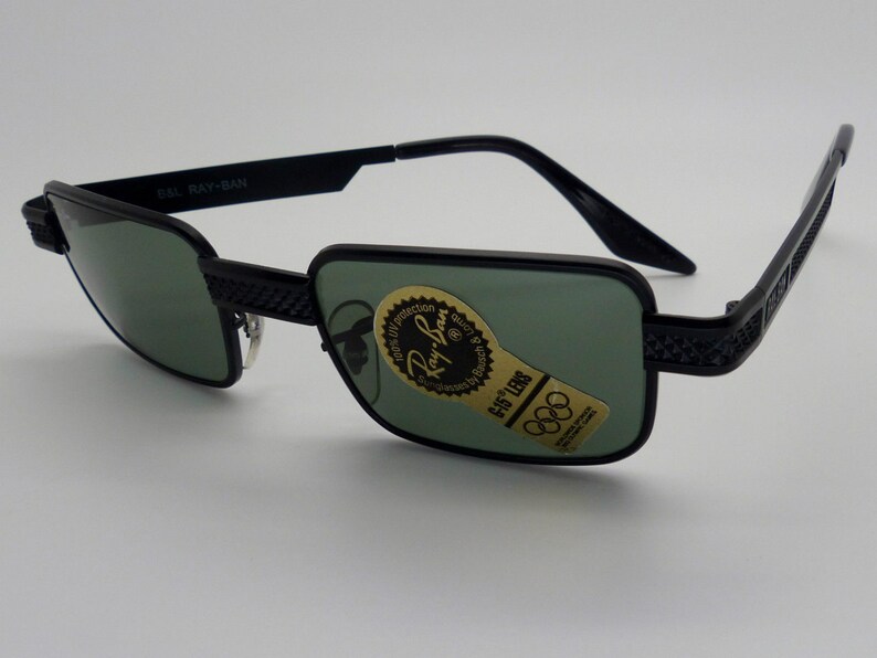 May include: Black Ray-Ban sunglasses with green lenses. The rectangular frames have a textured design on the temples and bridge. The lenses have a sticker with the Ray-Ban logo and Olympic rings. The text "G-15 LENS" is visible.