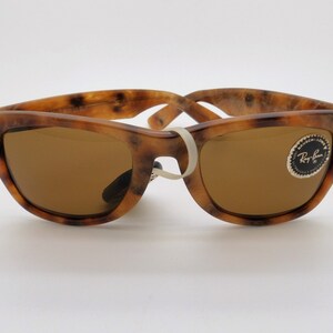 May include: Tortoiseshell-patterned sunglasses with brown lenses. The frames have a speckled brown and tan design. The sunglasses have a white band holding the arms together. A Ray-Ban logo sticker is on the right lens.