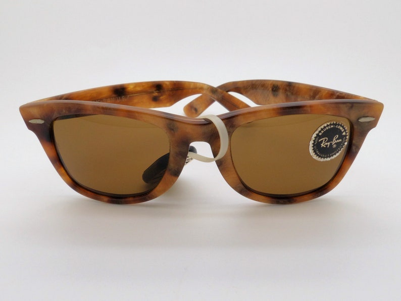 May include: Tortoiseshell-patterned sunglasses with brown lenses. The frames have a speckled brown and tan design. The sunglasses have a white band holding the arms together. A Ray-Ban logo sticker is on the right lens.