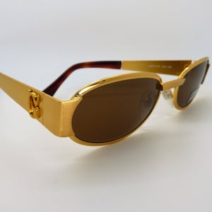 Puede incluir: Gafas de sol doradas con lentes marrones. Las monturas tienen forma rectangular con acabado cepillado. Las patillas son de color dorado macizo y las varillas son de color marrón oscuro. Diseño clásico y elegante.