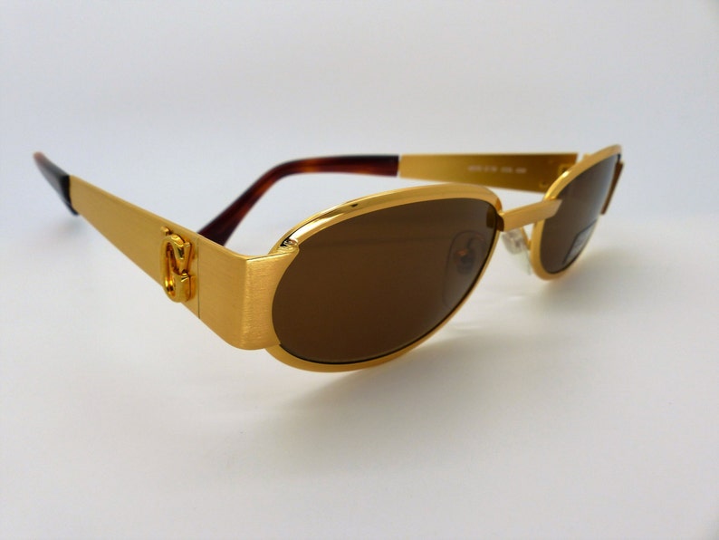 K&ouml;nnte beinhalten: Goldfarbene Sonnenbrille mit braunen Gl&auml;sern. Die Fassungen haben eine rechteckige Form mit geb&uuml;rsteter Oberfl&auml;che. Die B&uuml;gel sind goldfarben und die B&uuml;gel sind dunkelbraun. Die Sonnenbrille hat ein klassisches, elegantes Design.