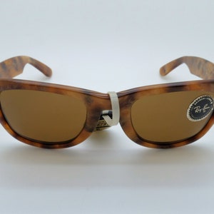 May include: Brown Wayfarer-style sunglasses with a tortoiseshell pattern on the frames. The lenses are a solid brown color. A white tag is attached to the bridge. The Ray-Ban logo is visible on the right lens.