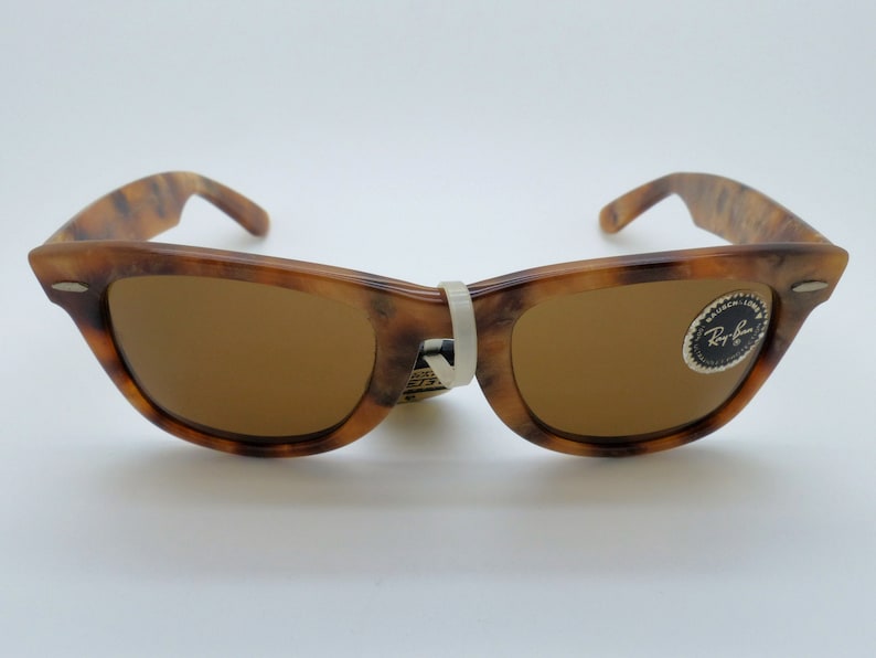 May include: Brown Wayfarer-style sunglasses with a tortoiseshell pattern on the frames. The lenses are a solid brown color. A white tag is attached to the bridge. The Ray-Ban logo is visible on the right lens.