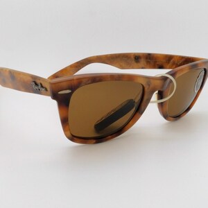 May include: Tortoiseshell-patterned sunglasses with brown lenses. The frames have a classic Wayfarer design, featuring a subtle logo on the temples. The lenses are a warm brown color, and the glasses are set against a white background.