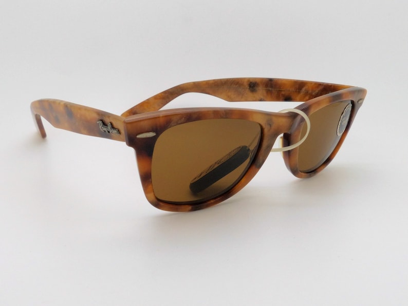 May include: Tortoiseshell-patterned sunglasses with brown lenses. The frames have a classic Wayfarer design, featuring a subtle logo on the temples. The lenses are a warm brown color, and the glasses are set against a white background.