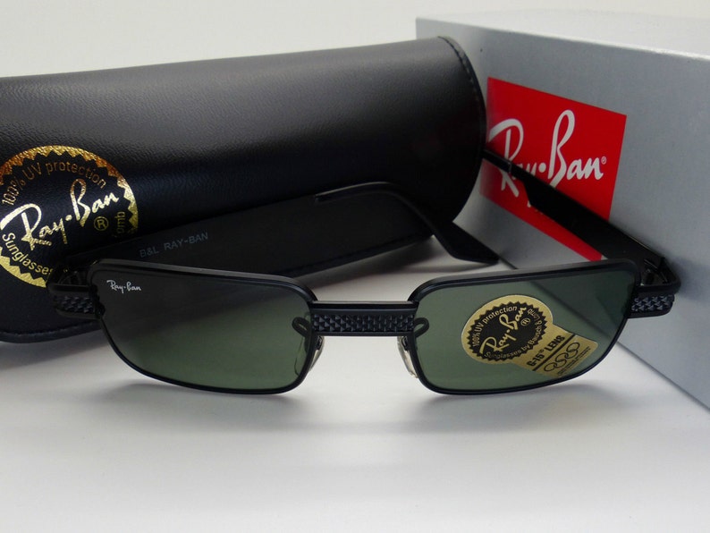 May include: Black Ray-Ban sunglasses with green lenses and a black frame. The sunglasses are displayed with a black case and a box with the Ray-Ban logo. The lenses have a sticker that says "100% UV protection".
