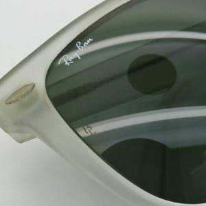 May include: Close-up of a pair of sunglasses with a silver frame and dark lenses. The Ray-Ban logo is visible on the lens. The sunglasses have a classic design.