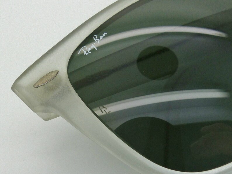 May include: Close-up of a pair of sunglasses with a silver frame and dark lenses. The Ray-Ban logo is visible on the lens. The sunglasses have a classic design.