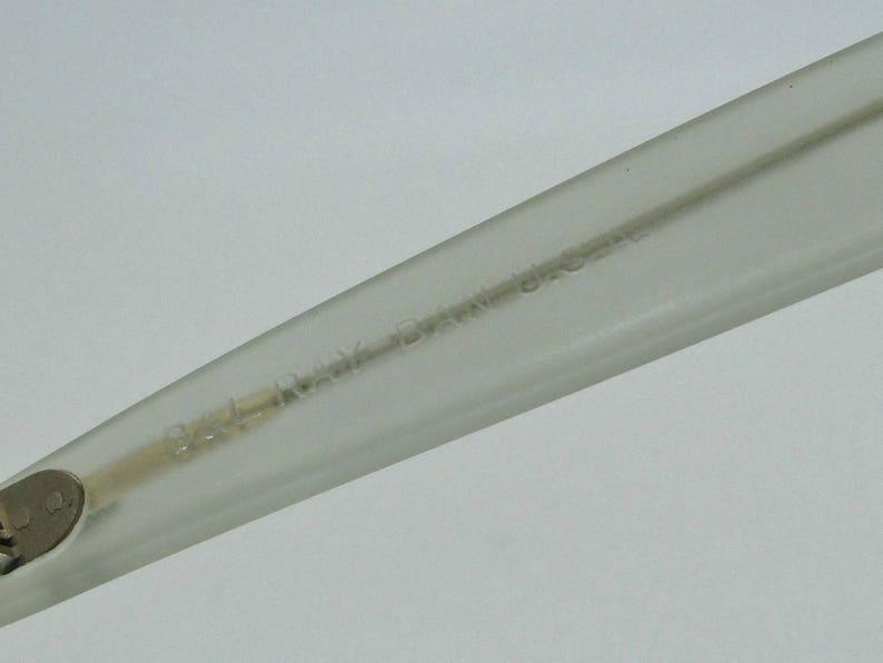 May include: Close-up of a clear eyeglass arm with the text "BAL RAY BRAZIL U.S.A." etched into the plastic. The arm is angled, showing the hinge and a portion of the frame.