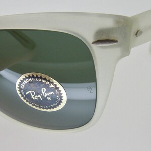 May include: Close-up of a pair of off-white Ray-Ban sunglasses. The lenses are a dark green color. The Ray-Ban logo is visible on the lens. The frame has a matte finish and a classic design.