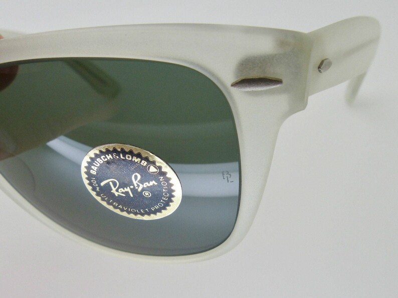 May include: Close-up of a pair of off-white Ray-Ban sunglasses. The lenses are a dark green color. The Ray-Ban logo is visible on the lens. The frame has a matte finish and a classic design.