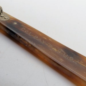 May include: Close-up of a vintage pair of sunglasses with a tortoiseshell pattern. The arm of the glasses is visible, with the text "B&L RAY-BAN U.S.A." engraved on the surface. The hinge is silver.