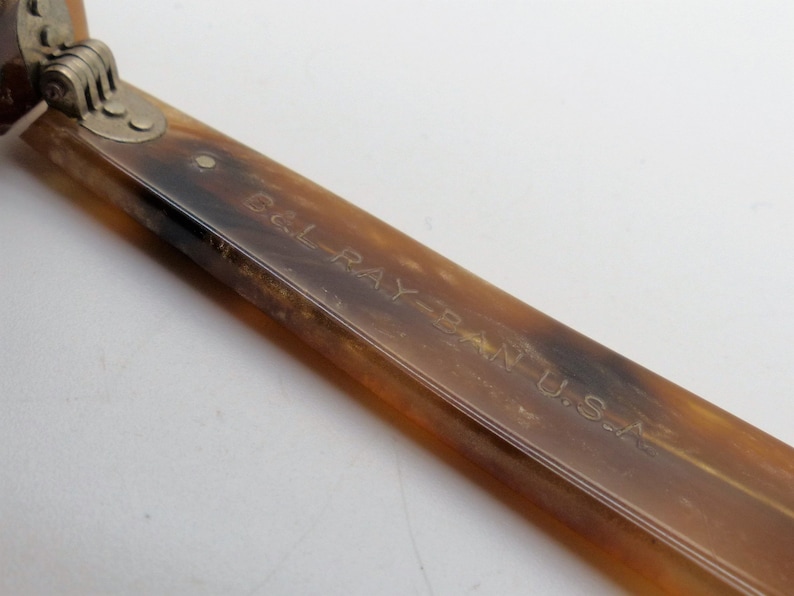 May include: Close-up of a vintage pair of sunglasses with a tortoiseshell pattern. The arm of the glasses is visible, with the text "B&L RAY-BAN U.S.A." engraved on the surface. The hinge is silver.