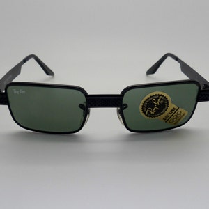 May include: Black Ray-Ban sunglasses with green lenses. The rectangular frames have a textured design on the temples and bridge. A sticker with the Ray-Ban logo and Olympic rings is on the lens.