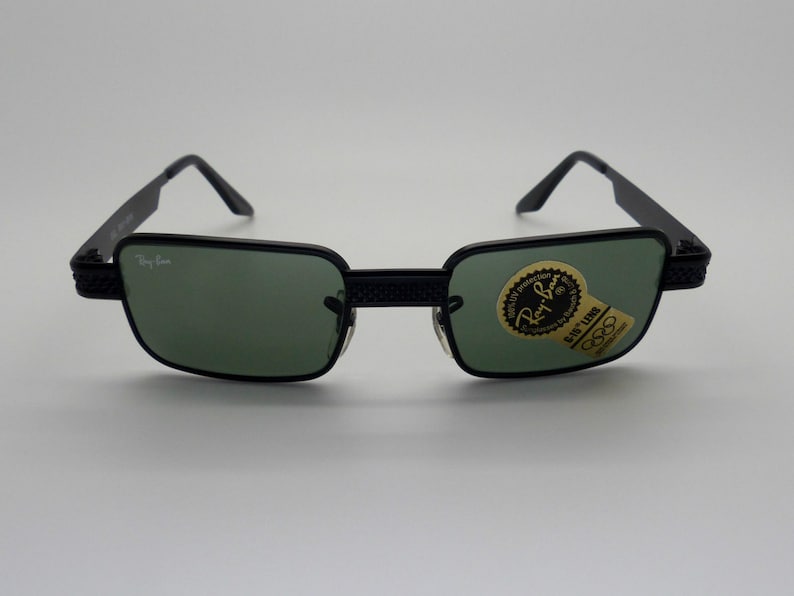 May include: Black Ray-Ban sunglasses with green lenses. The rectangular frames have a textured design on the temples and bridge. A sticker with the Ray-Ban logo and Olympic rings is on the lens.