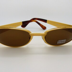 K&ouml;nnte beinhalten: Goldfarbene Sonnenbrille mit braunen Gl&auml;sern. Die Sonnenbrille hat eine rechteckige Form mit einem goldenen Steg und B&uuml;geln. Die B&uuml;gel haben ein Schildpattmuster. Die Gl&auml;ser haben einen Aufkleber mit der Aufschrift "VERSACE".