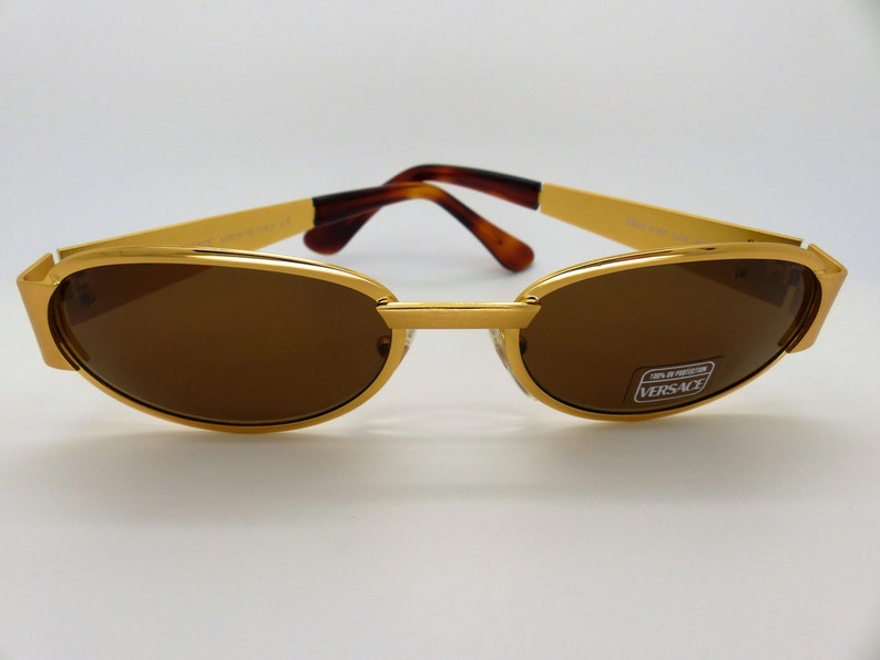 K&ouml;nnte beinhalten: Goldfarbene Sonnenbrille mit braunen Gl&auml;sern. Die Sonnenbrille hat eine rechteckige Form mit einem goldenen Steg und B&uuml;geln. Die B&uuml;gel haben ein Schildpattmuster. Die Gl&auml;ser haben einen Aufkleber mit der Aufschrift "VERSACE".