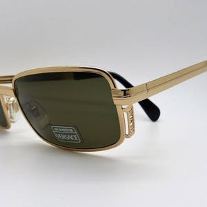 Puede incluir: Gafas de sol con montura dorada y lentes oscuras. Las monturas rectangulares tienen un acabado dorado con patillas negras. Las lentes tienen un tinte verde. Las gafas de sol tienen una pegatina de Versace en la lente.