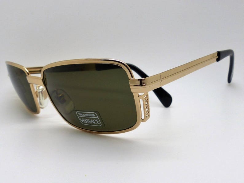 Puede incluir: Gafas de sol con montura dorada y lentes oscuras. Las monturas rectangulares tienen un acabado dorado con patillas negras. Las lentes tienen un tinte verde. Las gafas de sol tienen una pegatina de Versace en la lente.