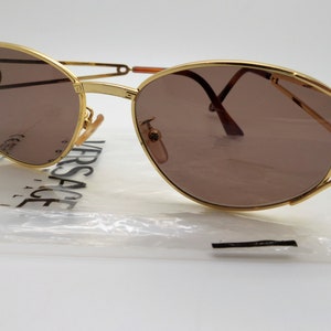 Puede incluir: Gafas de sol doradas con lentes marrones. Las gafas tienen un diseño distintivo con una montura dorada y un puente único. Las lentes son de forma ovalada. El nombre de la marca "VERSACE" es visible en el embalaje.