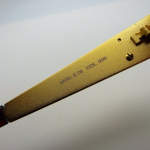 K&ouml;nnte beinhalten: Nahaufnahme eines goldfarbenen Brillenb&uuml;gels. Auf dem B&uuml;gel steht "MOD. S 78 COL. 030". Der Rahmen ist mit einem braunen B&uuml;gel verbunden. Das Scharnier ist sichtbar.