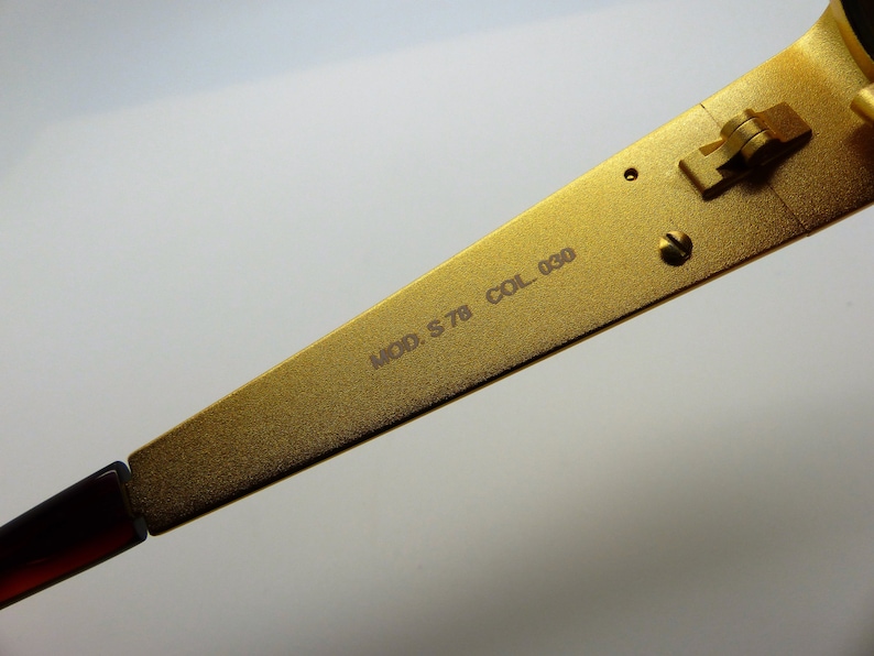 K&ouml;nnte beinhalten: Nahaufnahme eines goldfarbenen Brillenb&uuml;gels. Auf dem B&uuml;gel steht "MOD. S 78 COL. 030". Der Rahmen ist mit einem braunen B&uuml;gel verbunden. Das Scharnier ist sichtbar.