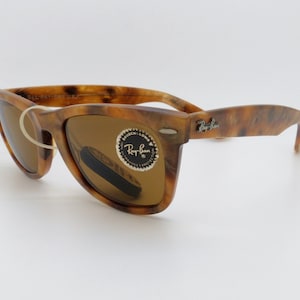 May include: Tortoiseshell-patterned sunglasses with brown lenses. The sunglasses have a classic Wayfarer style with the Ray-Ban logo on the temple. The lenses have a protective sticker. The frame is a mix of brown and amber.