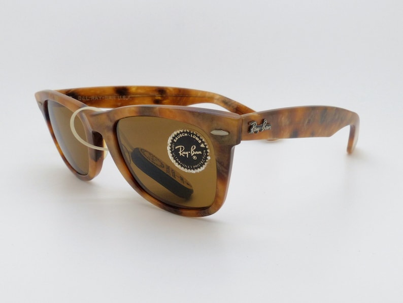 May include: Tortoiseshell-patterned sunglasses with brown lenses. The sunglasses have a classic Wayfarer style with the Ray-Ban logo on the temple. The lenses have a protective sticker. The frame is a mix of brown and amber.