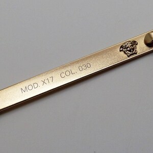 Puede incluir: Primer plano de una varilla de montura de gafas dorada. La varilla presenta el texto "MOD. X17 COL. 030" y un emblema de cabeza de Medusa. La varilla tiene una punta negra y una bisagra dorada.